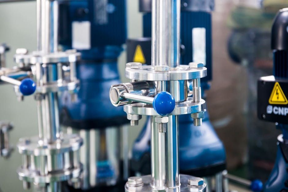Stainless steel lab valves and tubing with blue knobs in a blurred industrial setting