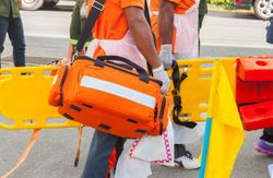 Person carrying an orange medical bag, near a stretcher and other emergency equipment.