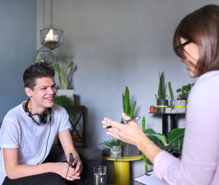 A person smiles while speaking with someone in a room with plants. They both sit on chairs.