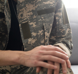 Person in camouflage uniform, hands clasped, resting on knees.