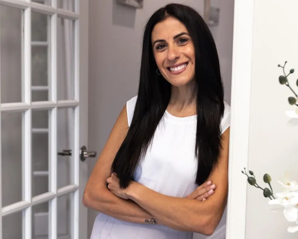 Woman with long dark hair smiles, arms crossed, leaning against a white doorway, bright setting.