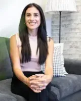 Woman seated on a gray sofa, smiling. She is wearing a light pink top and black pants. Living room setting.
