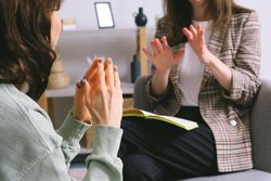 Two women in a therapy session; one gestures, the other clasps hands, with a notebook in front of them.