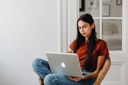 Woman sits in a wicker chair, looking at laptop. She wears a red shirt, jeans, and sits near a white door.