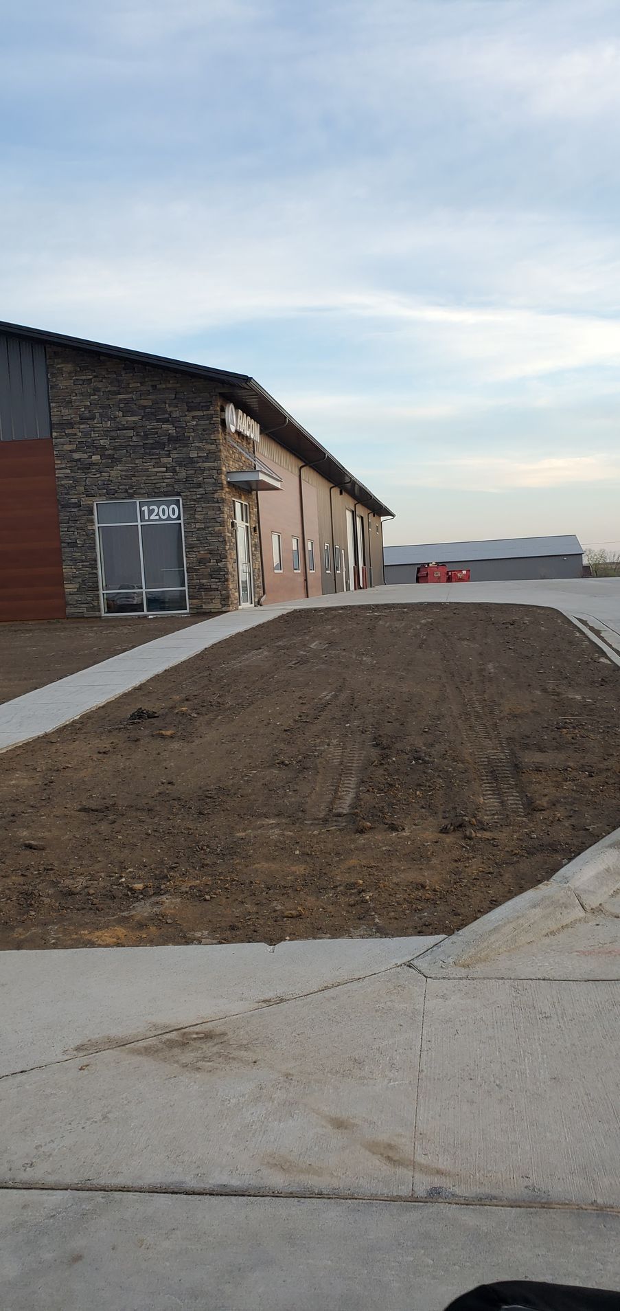 Building exterior with a stone facade, a paved path, and bare earth. Blue sky in the background.
