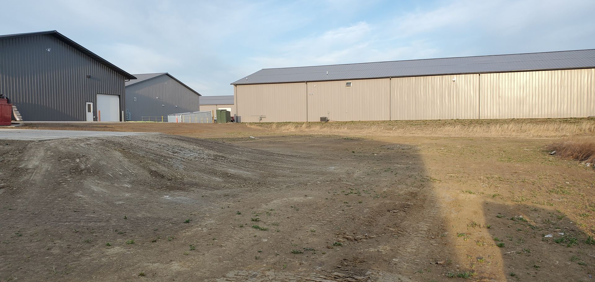 A wide shot of several industrial buildings with metal siding on a dirt lot. The sky is partly cloudy.