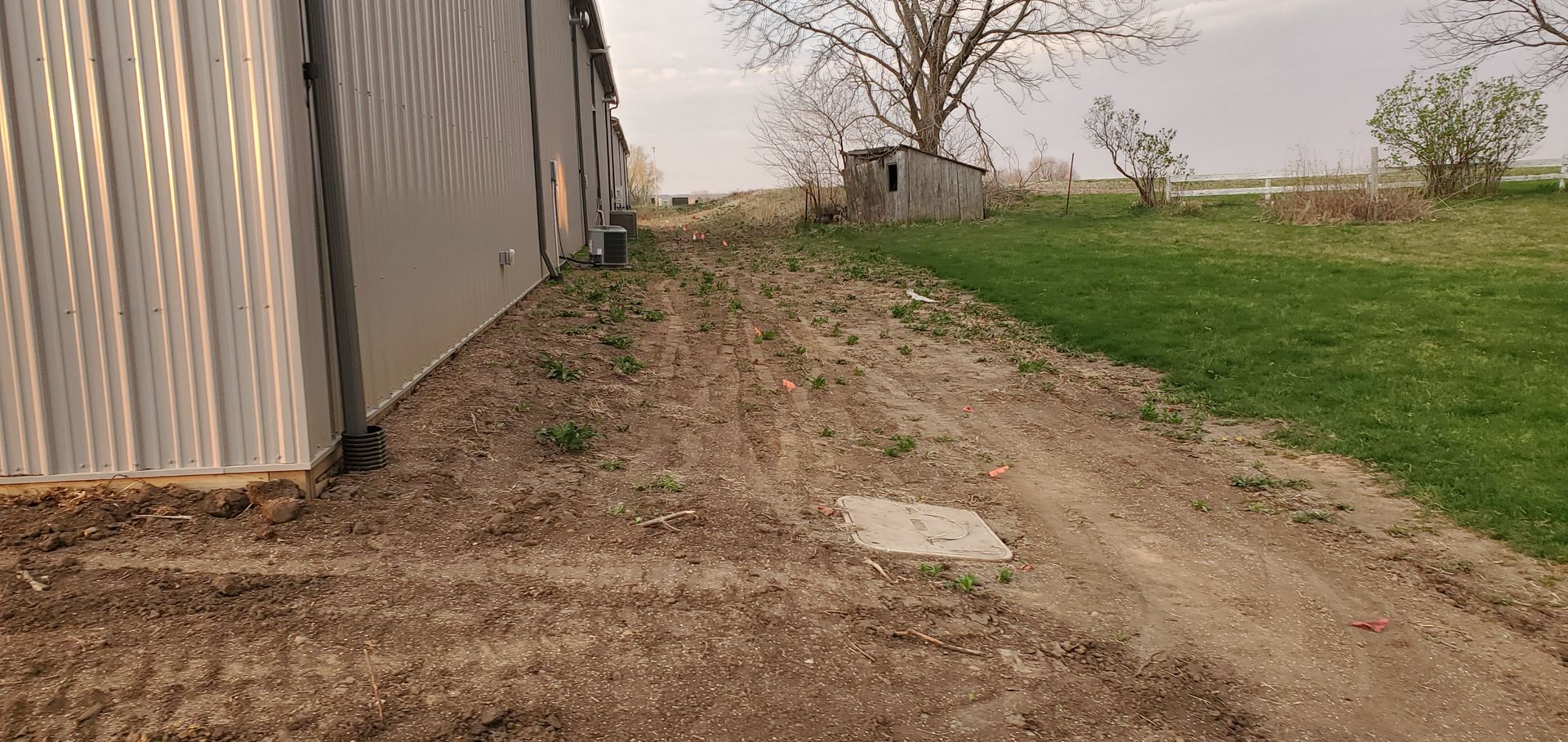 Dirt path beside a metal building, leading toward a grassy area with a small shed and trees.