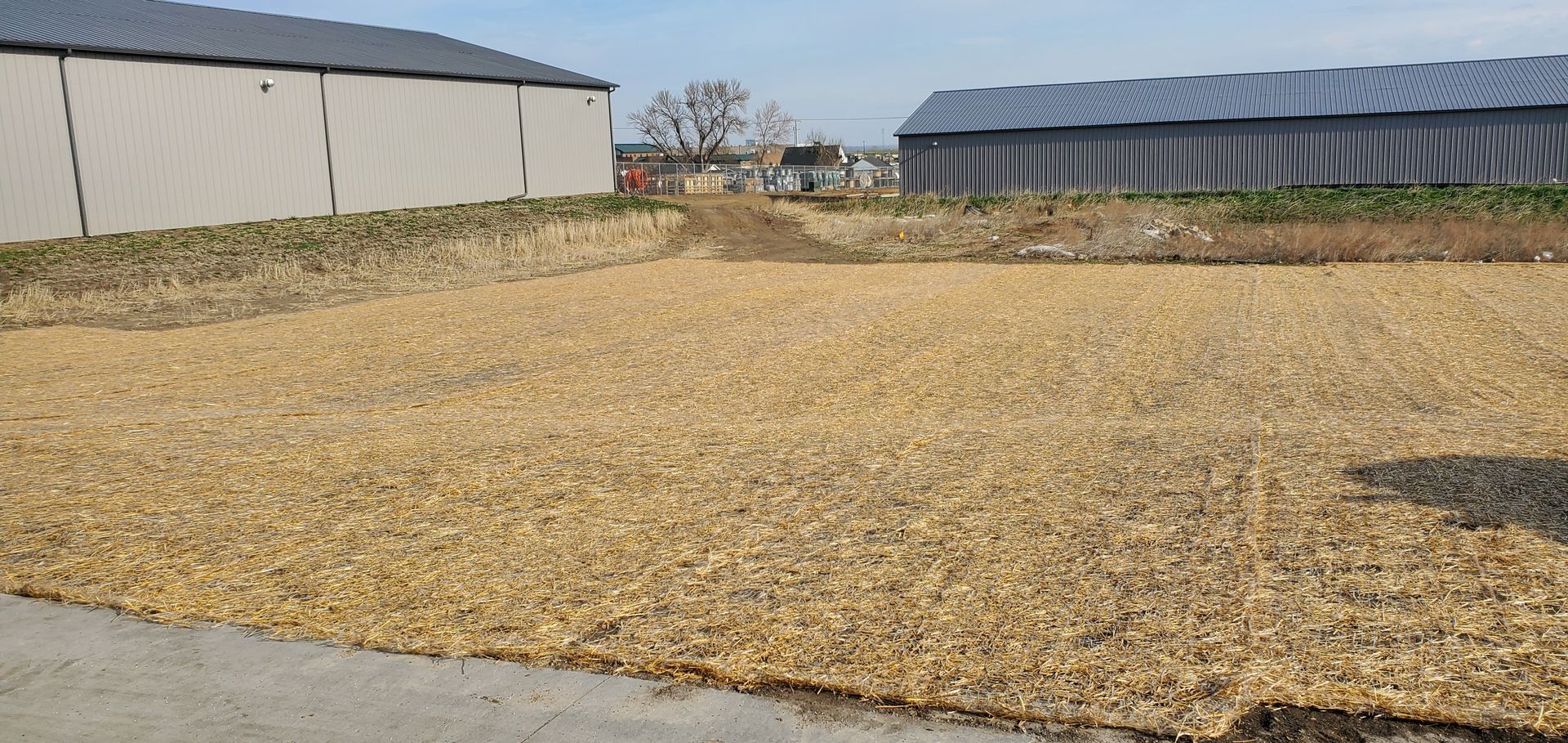 A field covered in tan-colored mulch with two large buildings in the background. 