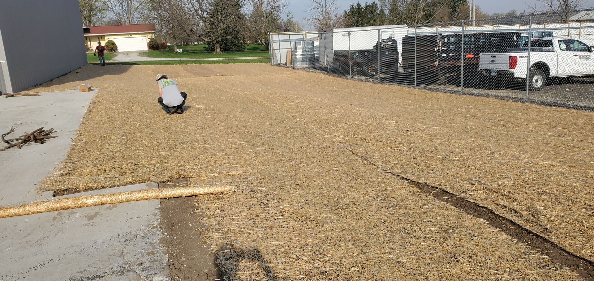 Person kneeling on wood chip ground.  Building, chain link fence, and parked vehicles in the background.