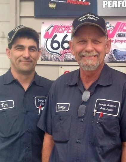 Two mechanics in dark blue uniforms stand together in front of a Jasper Engines sign and a Route 66 wall display.