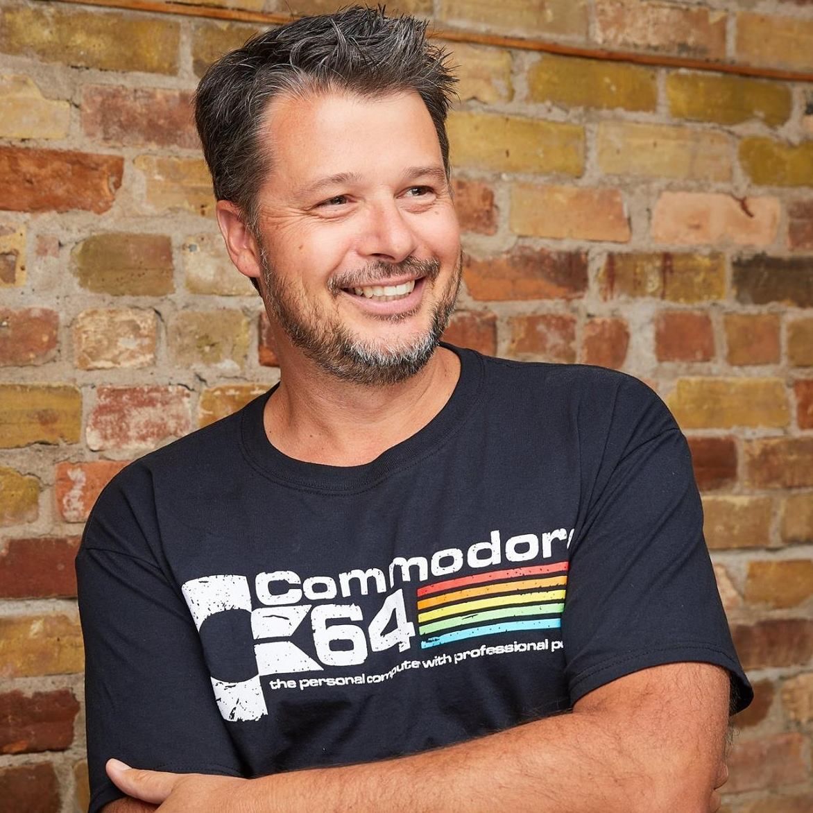 Man wearing a Commodore 64 t-shirt smiles against a brick wall background.