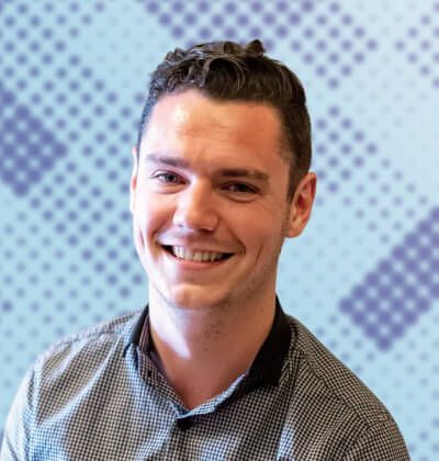 Man with dark hair smiling at the camera, wearing a gray shirt, against a blue and white patterned background.