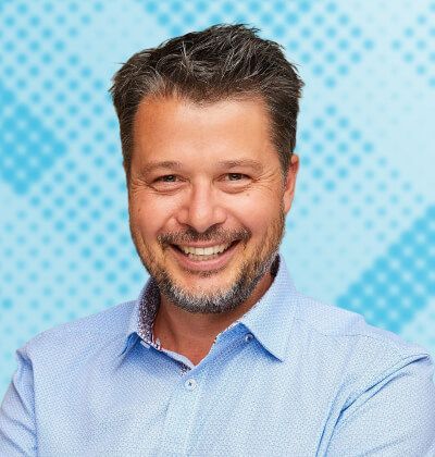 Smiling man with short hair and a graying beard, wearing a blue patterned shirt.
