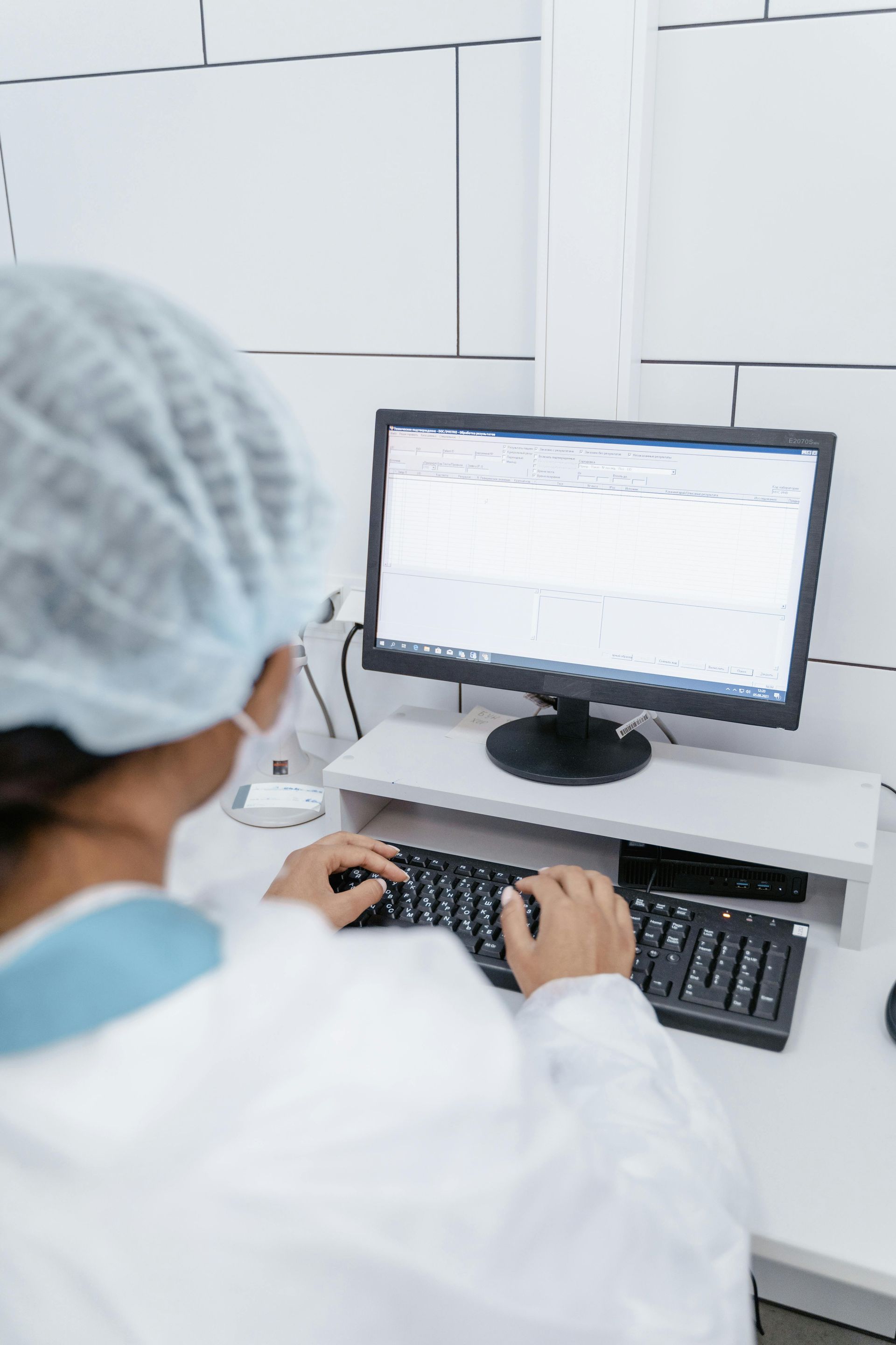 Person in lab coat and hairnet using a computer, typing on a keyboard, in a white-walled room.