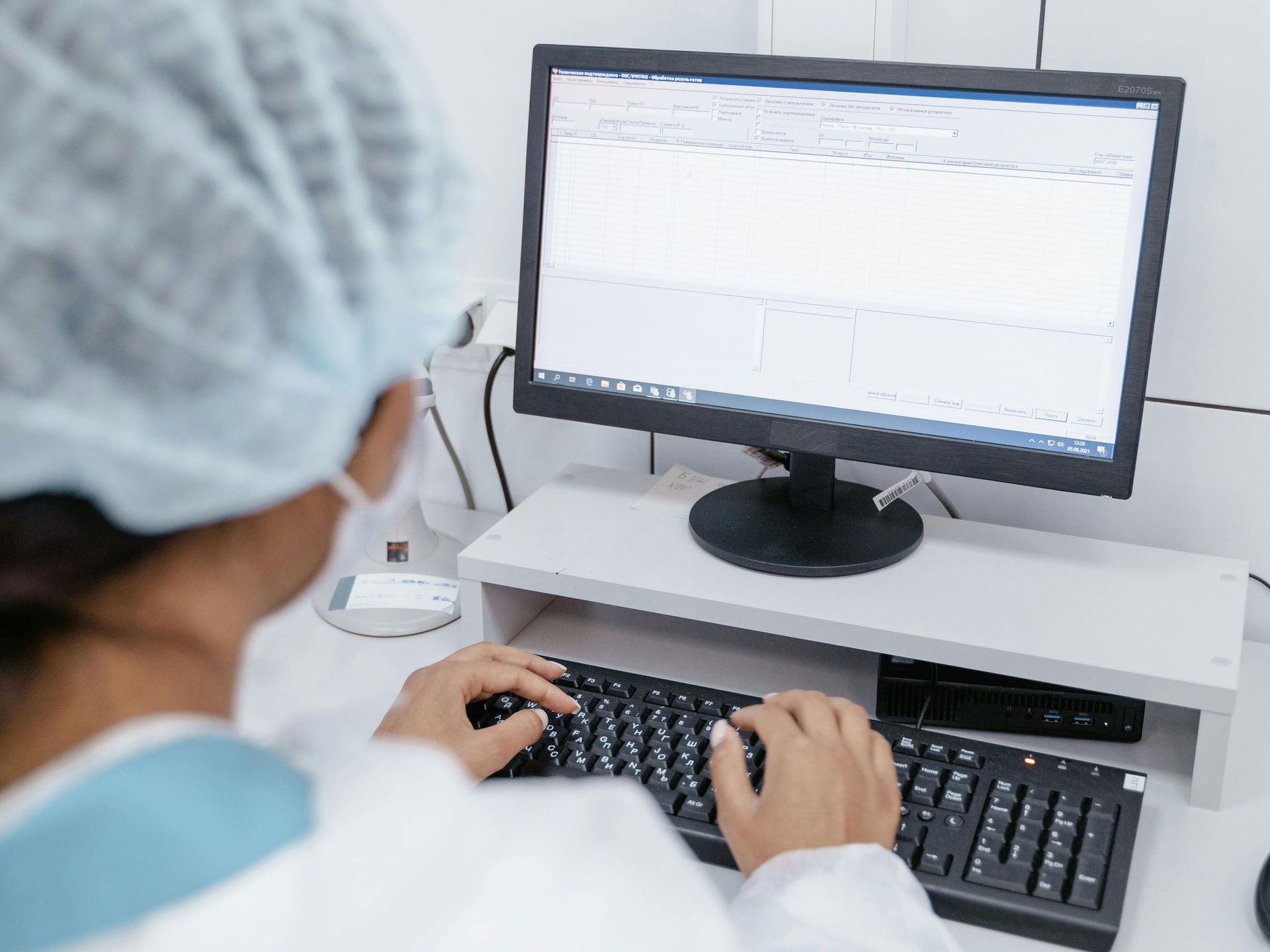 Person wearing a medical cap and mask typing on a computer at a desk.