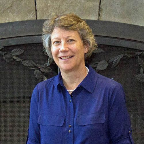 A woman in a blue shirt is smiling in front of a fireplace.