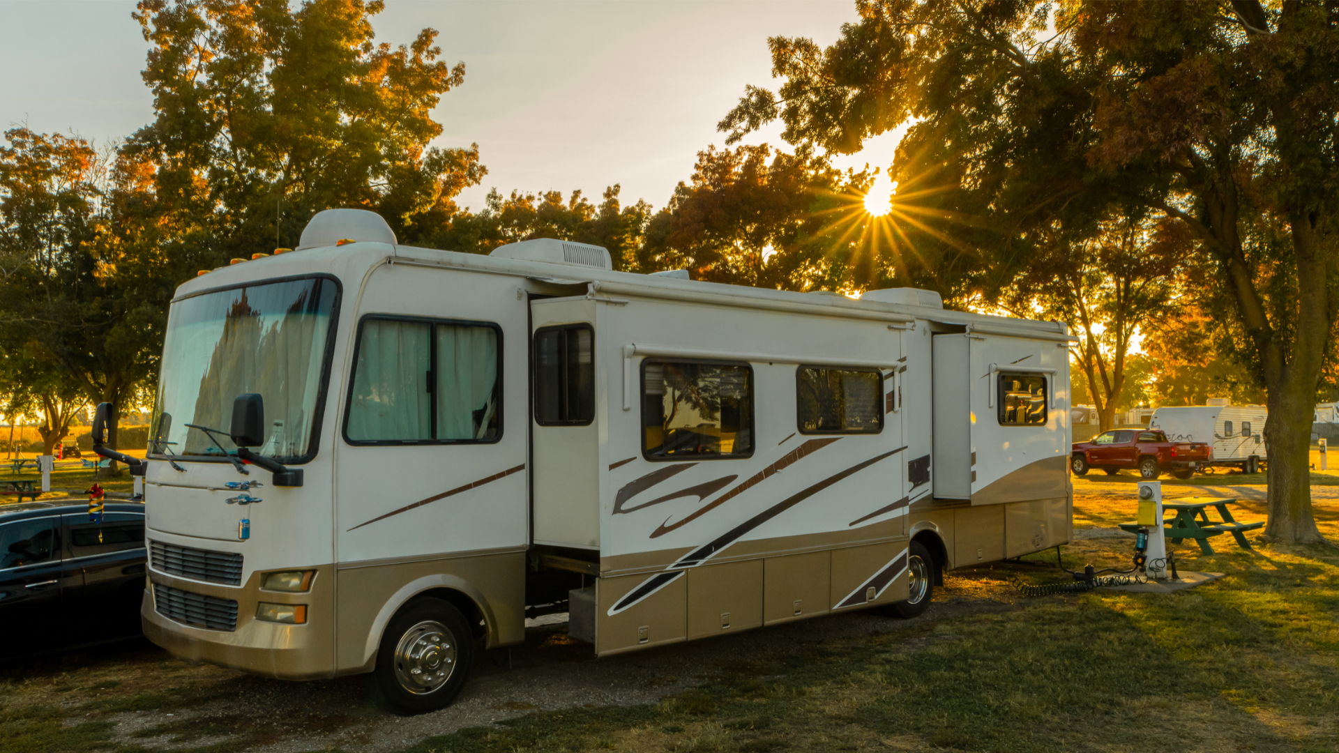 RV parked in a campground at sunset. Beige and white RV has an open side, with the sun shining through trees in the background.