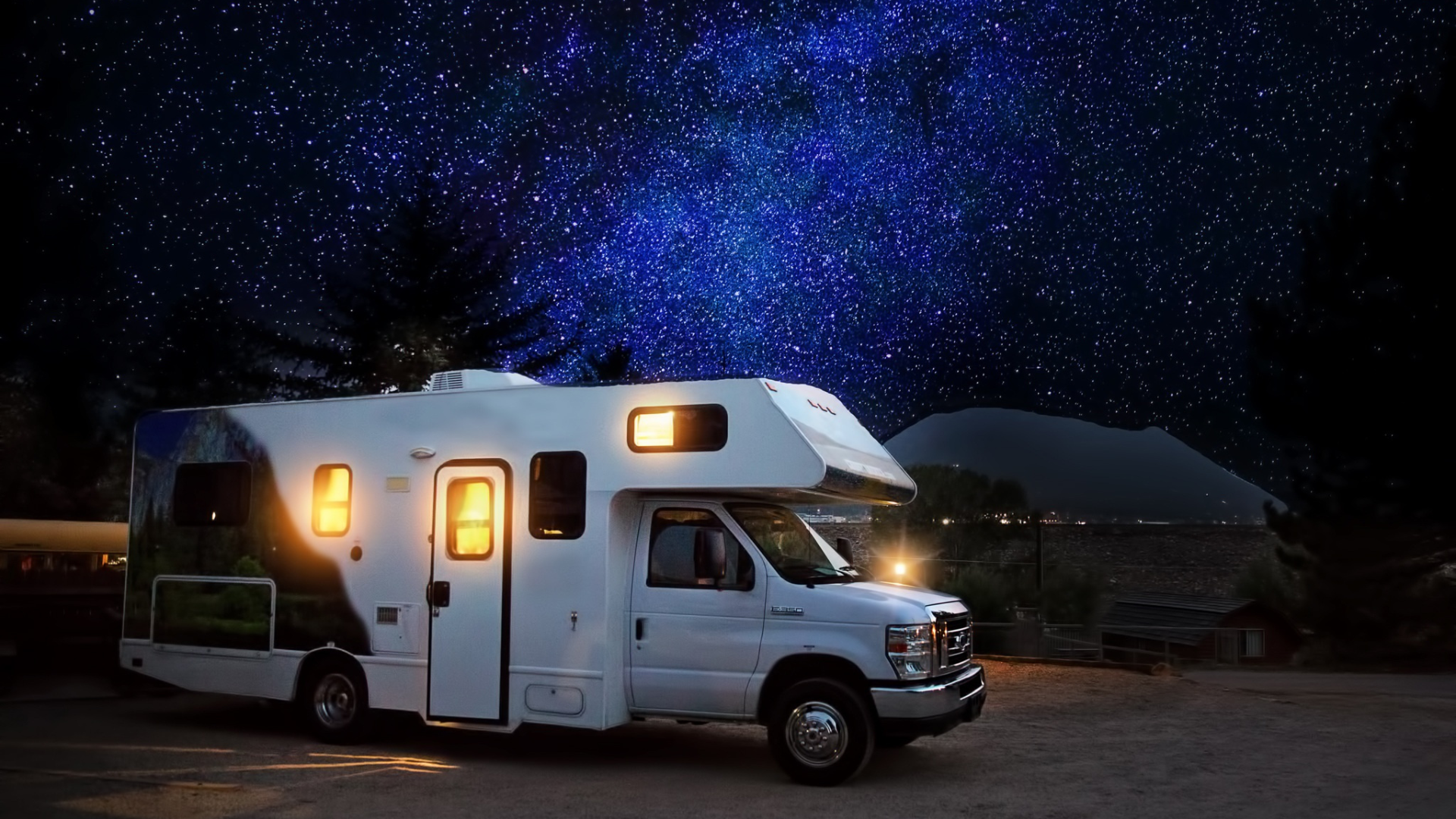 White RV parked under a dark night sky filled with stars. The RV's interior lights are on, casting a warm glow.