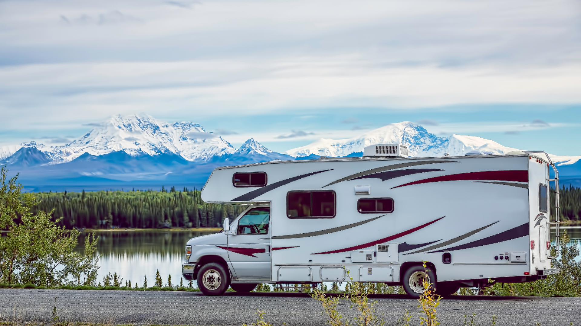 White RV parked on a road next to a lake, mountains with snowy peaks in the background.