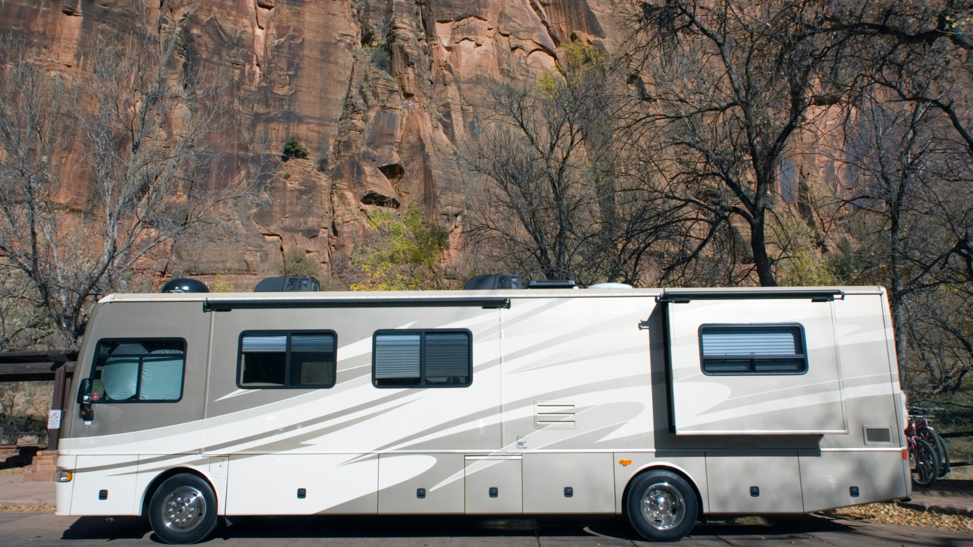 A large RV parked on asphalt in front of a rocky cliff. The RV is beige and has a slide-out.