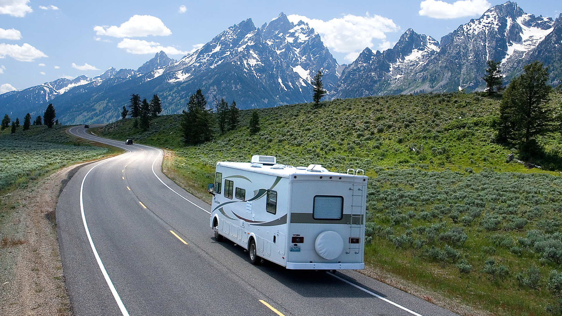 RV driving along a winding road through a mountain landscape. The sky is blue with some clouds.