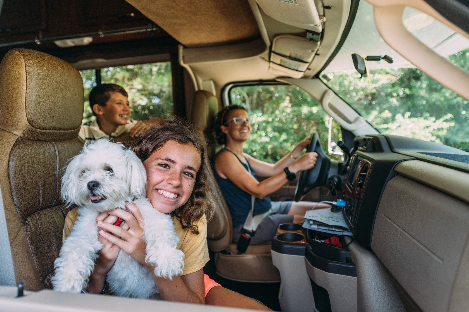 A family, including a child holding a white dog, smiles in the front of an RV while the mother drives.