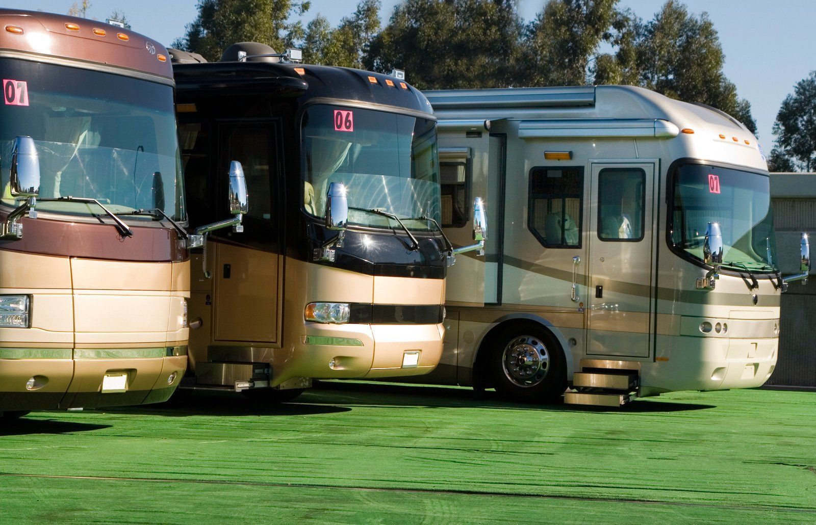 Three recreational vehicles parked on green grass under a sunny sky. They are various shades of gold, brown, and white.