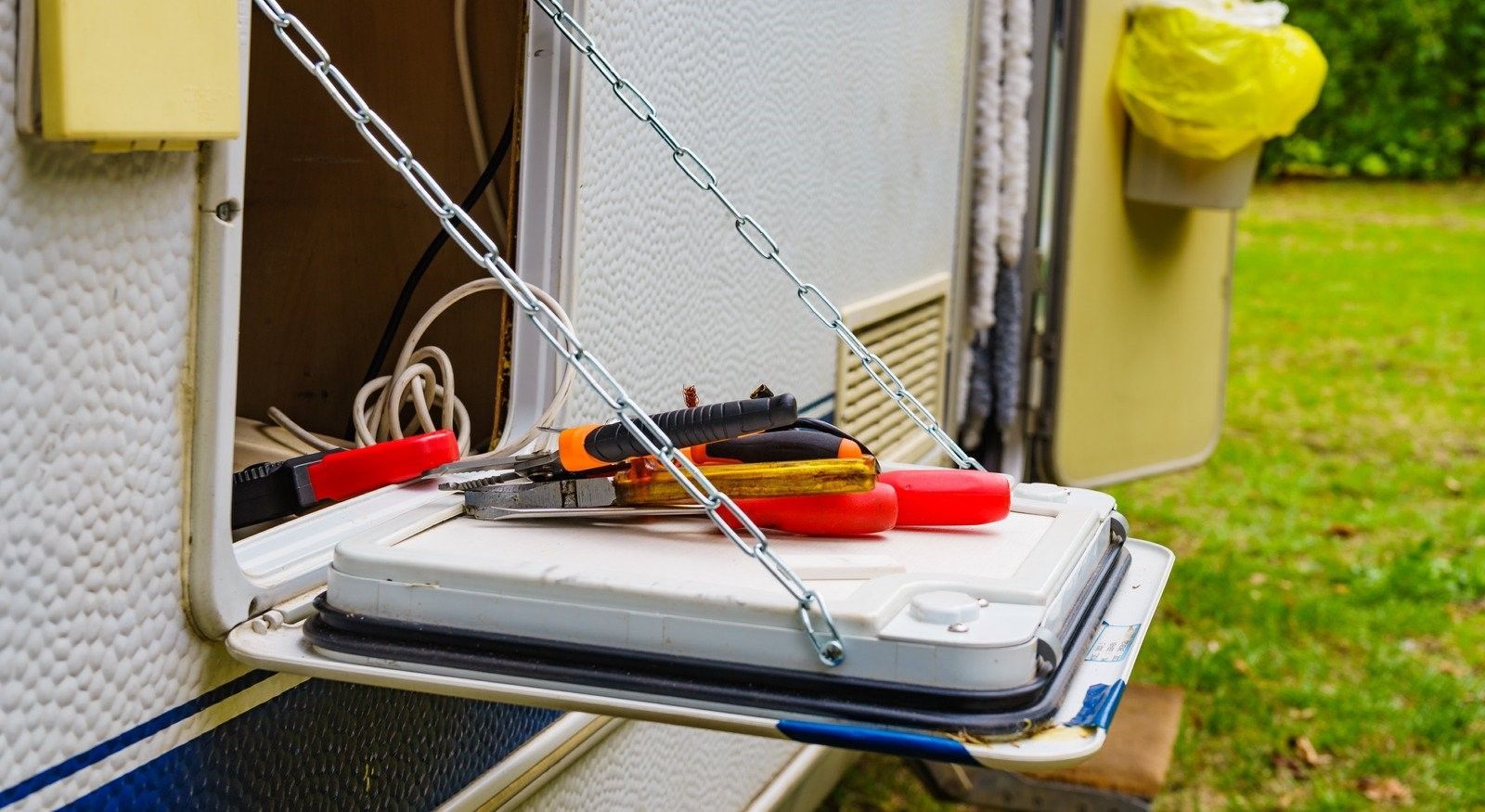A white, folding table is attached to the side of a camper. The table holds tools, and is supported by metal chains.