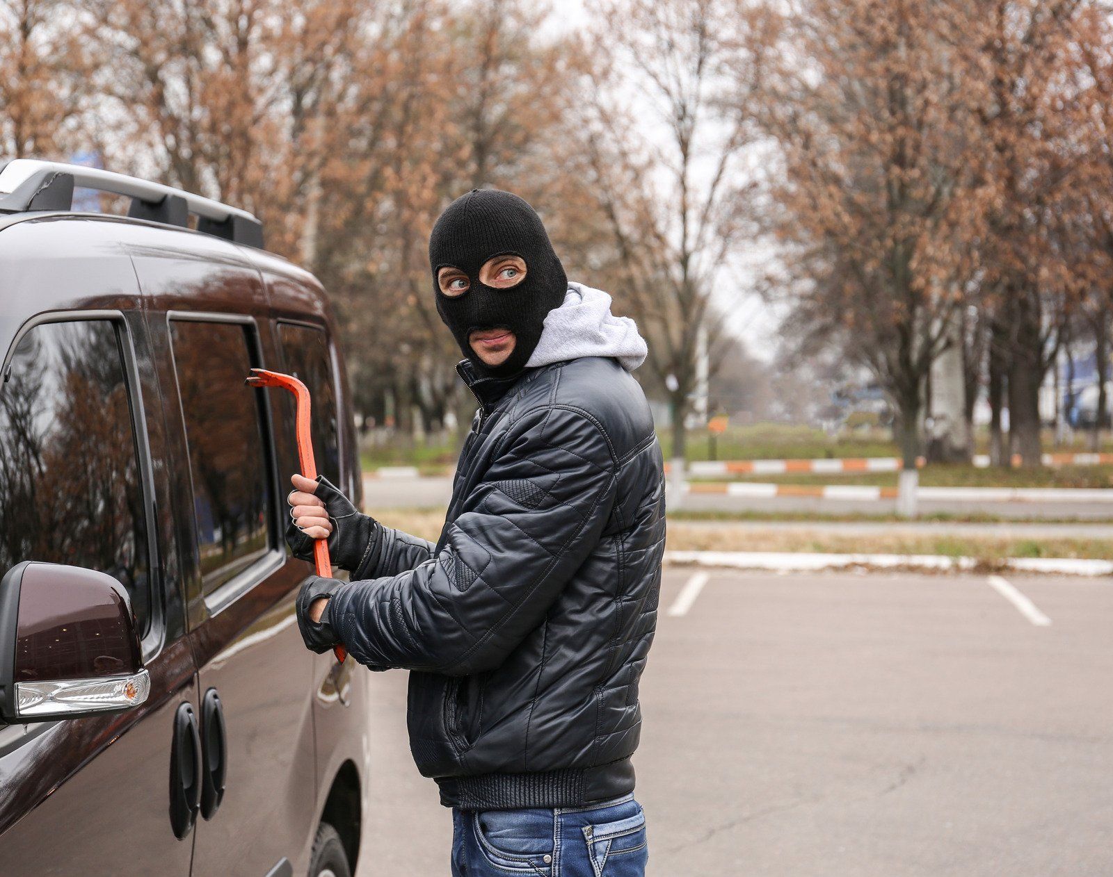 Person in a ski mask using a crowbar to break into a car window in a parking lot.