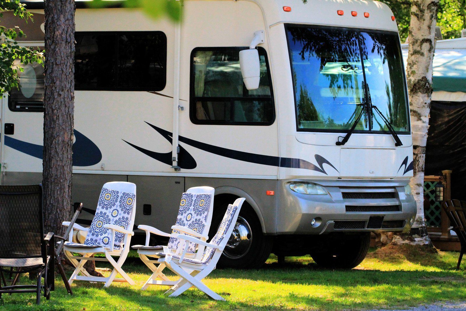 RV parked on green grass, with two white chairs in front. The setting is a campsite, with trees in the background.