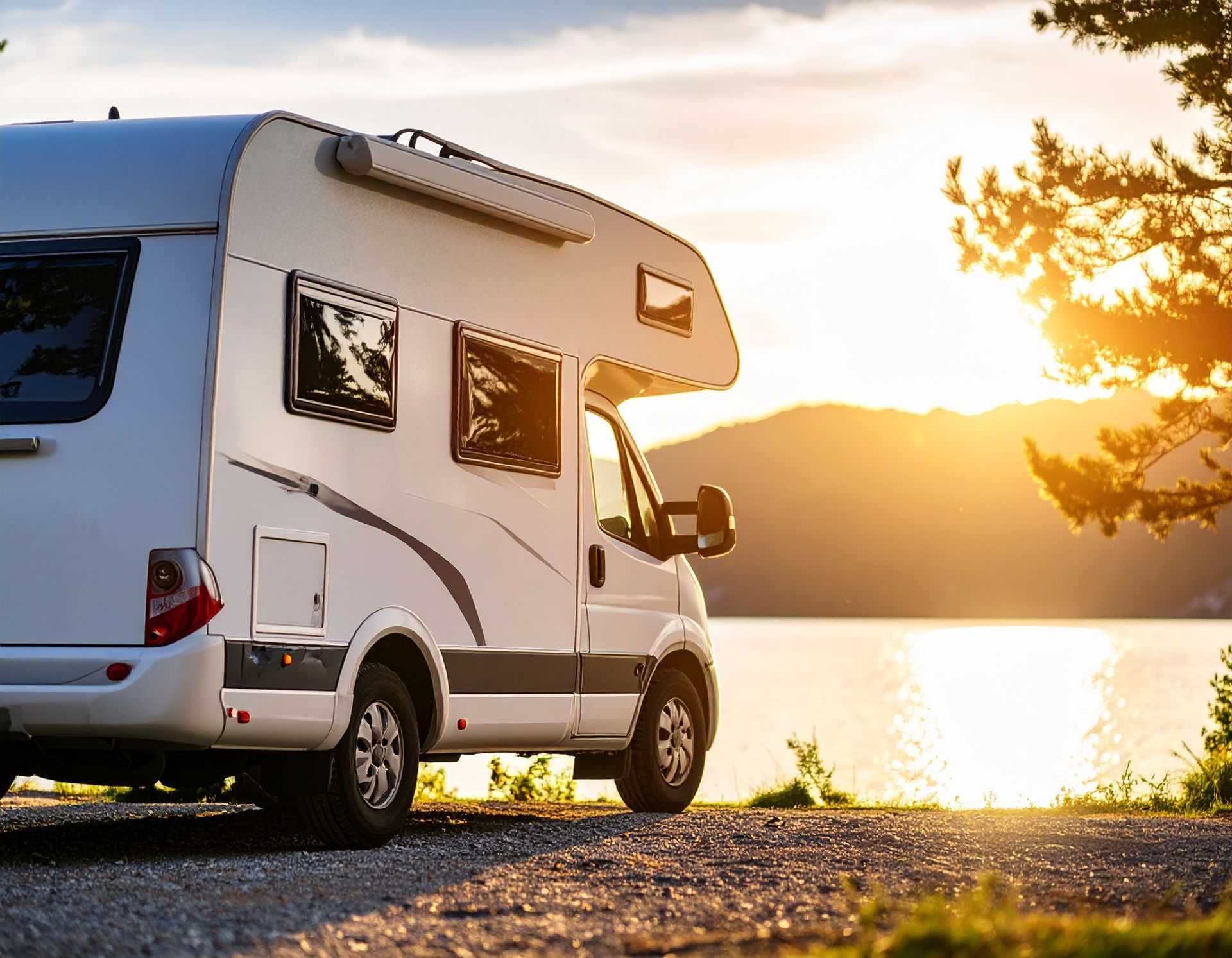 White RV parked near a lake at sunset; sun is setting behind the mountains, casting a golden glow.