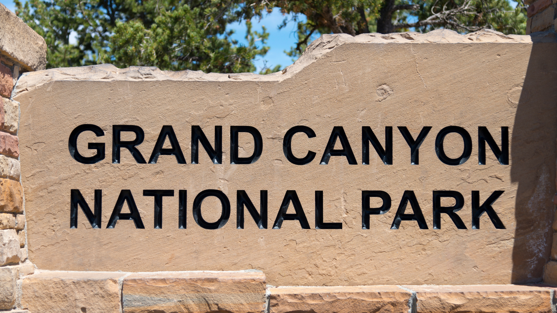 Grand Canyon National Park sign, tan stone with black text, set against a blue sky and green trees.