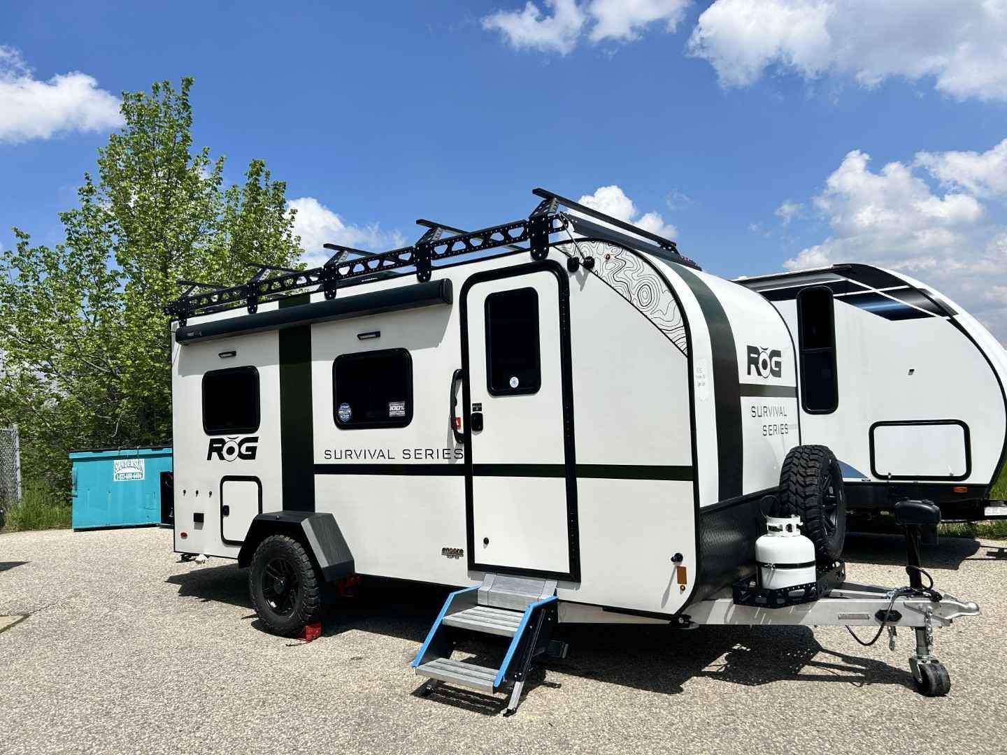 White and black R-Pod travel trailer with black roof rack, parked on gravel. Features a door with stairs and a spare tire.