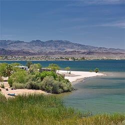 A scenic view of a lake with a sandy beach, green vegetation, and mountains in the background under a blue sky.