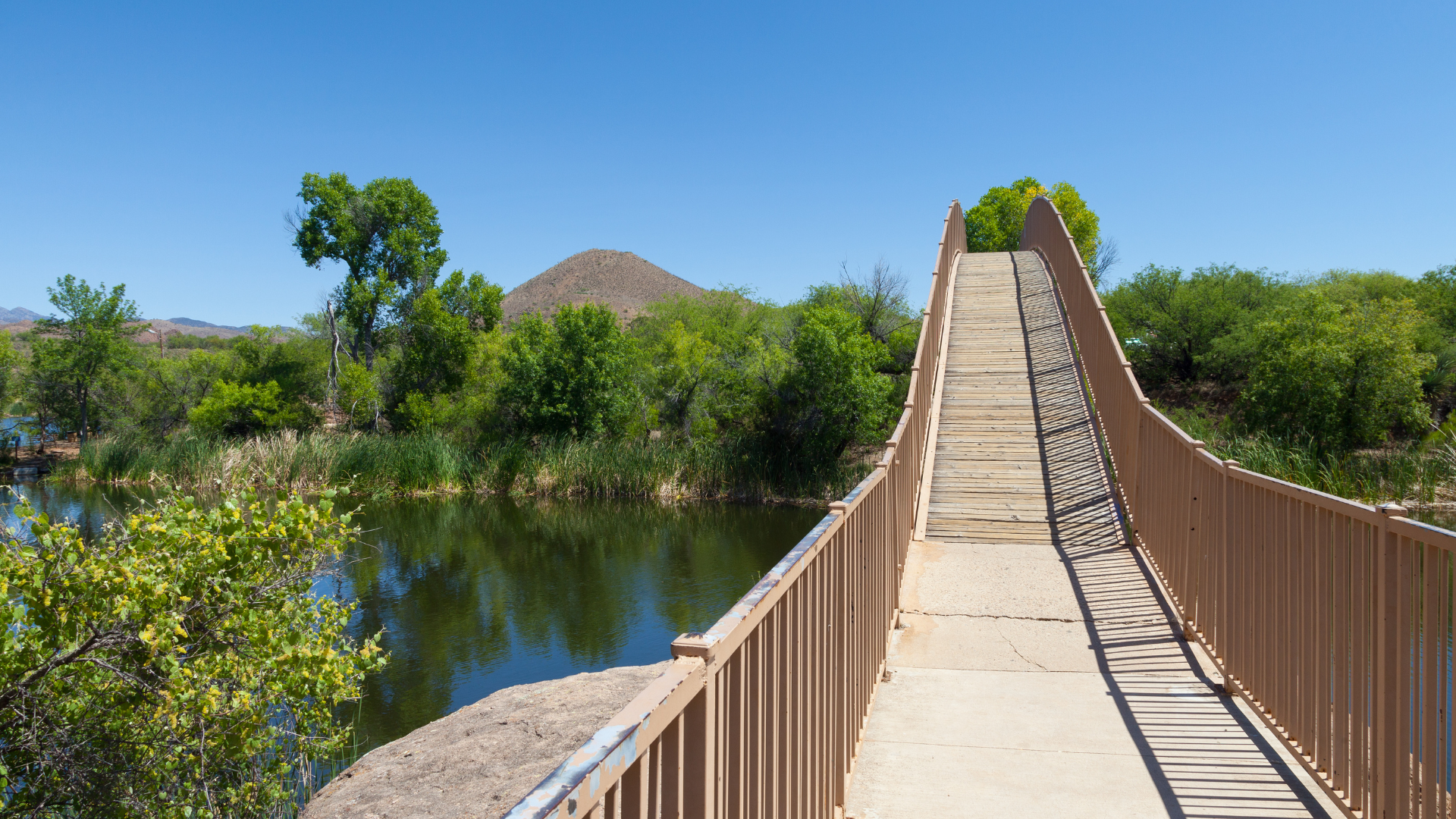 A brown suspension bridge over a calm lake leads to a green treeline and a mountain under a blue sky.