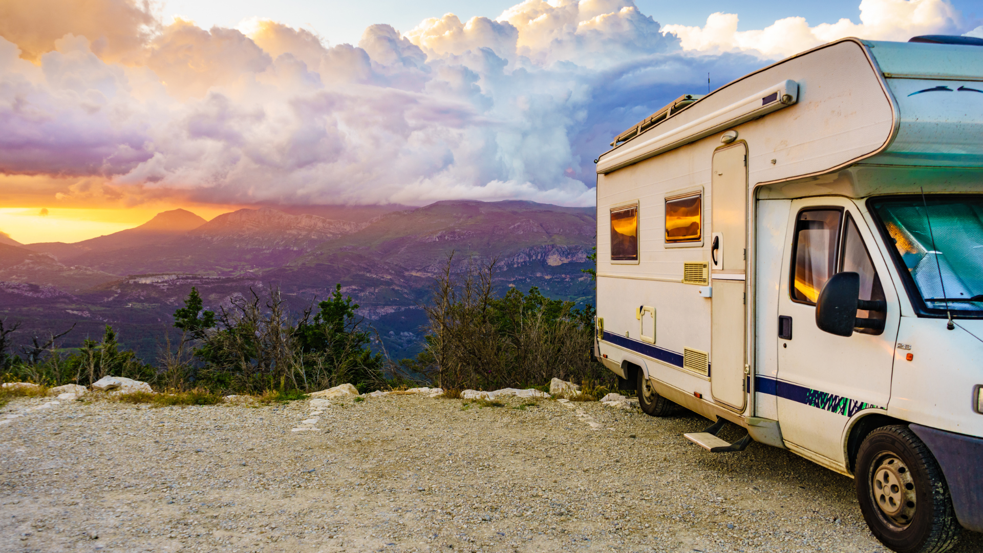 RV parked on a gravel hilltop overlooking a landscape at sunset. Orange and yellow hues in the sky, green hills in the distance.