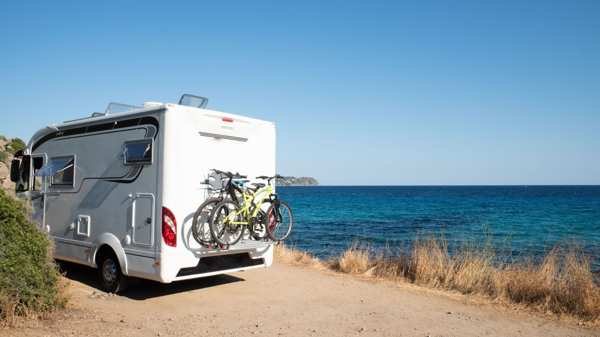 White RV parked on a dirt road next to a blue ocean. Two bicycles are mounted on the back.