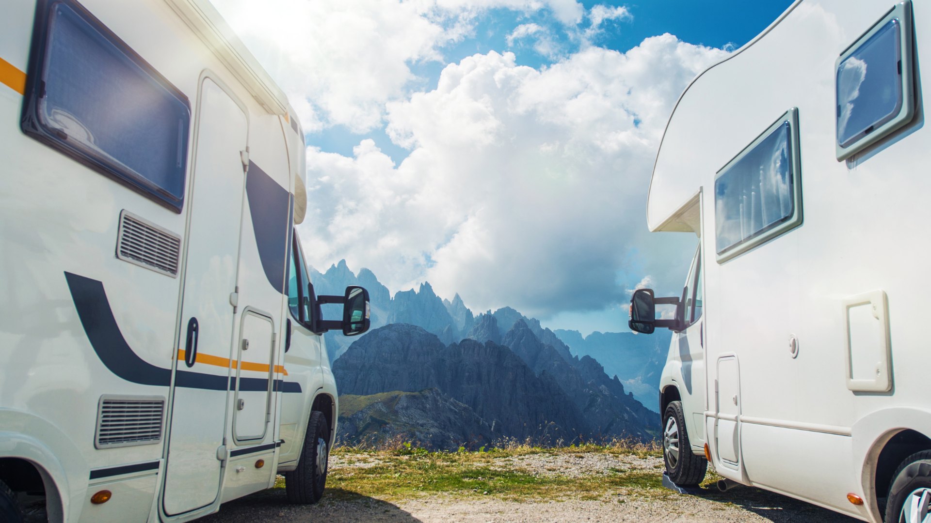 Two white RVs parked with mountain range backdrop on a sunny day.