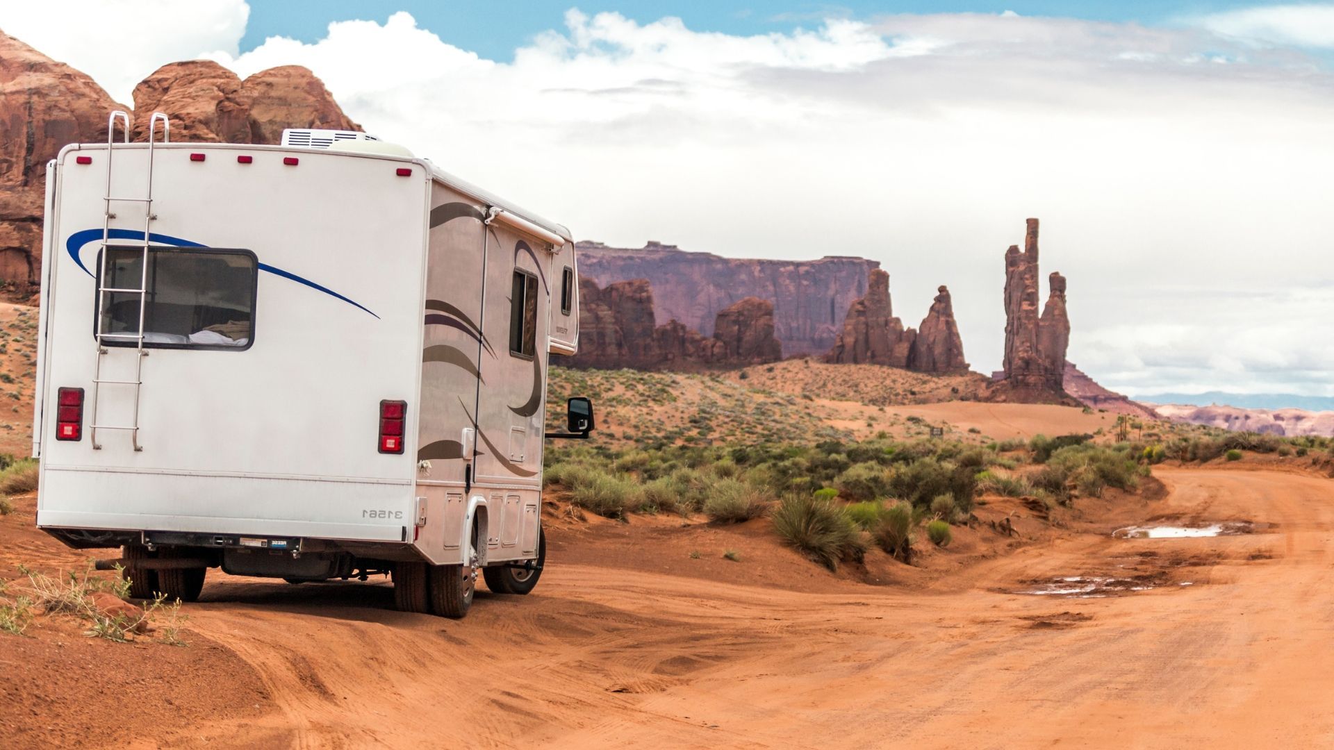 White RV parked on a dirt road in a desert landscape with red rock formations under a cloudy sky.