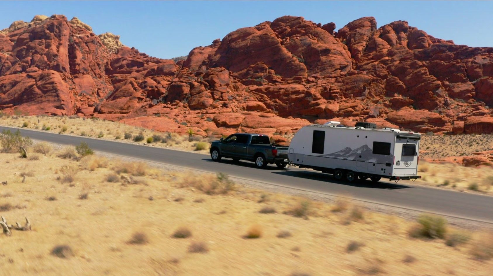 A truck towing a camper on a desert road with red rock formations in the background under a clear blue sky.