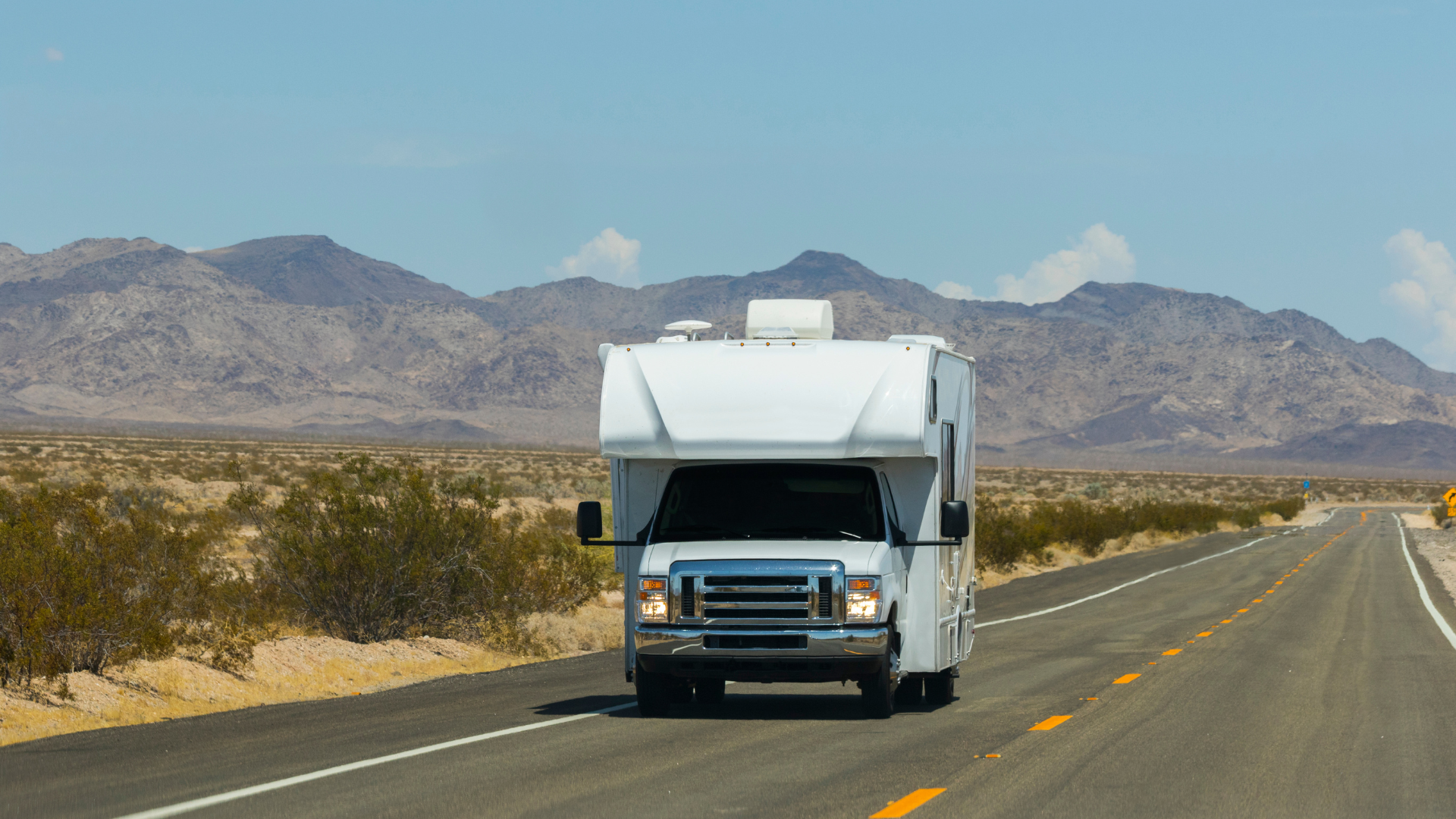 White RV traveling on a desert highway, with mountains in the background. Sunny day.