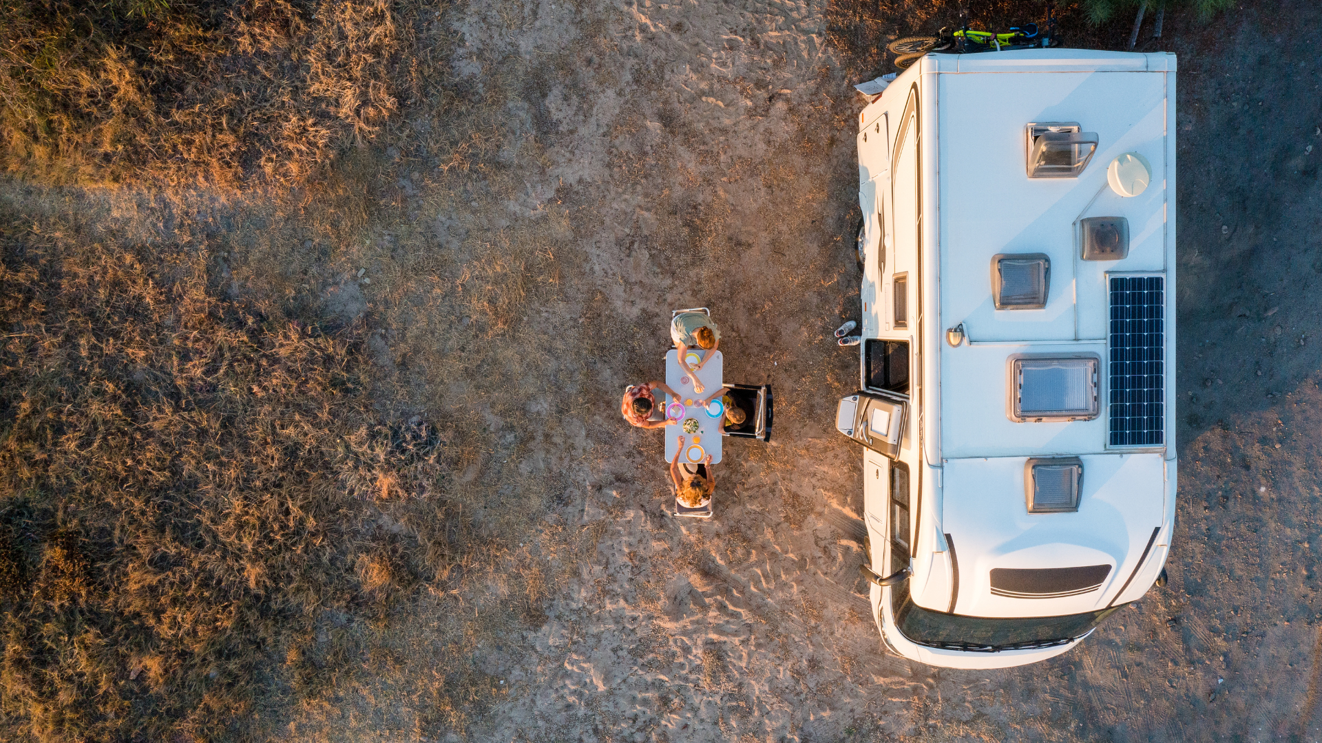 Overhead view of a white RV parked in a dirt area with a person at a table. Sunlight bathes the scene in warm hues.