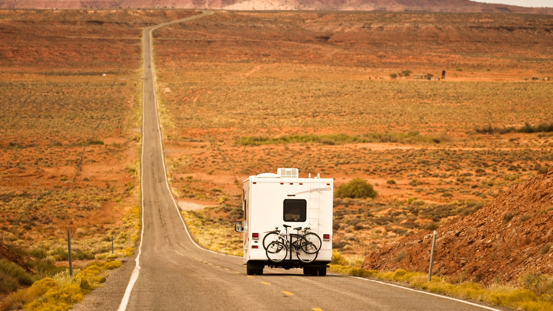 A white RV travels down a long, empty desert road, surrounded by reddish-brown terrain.