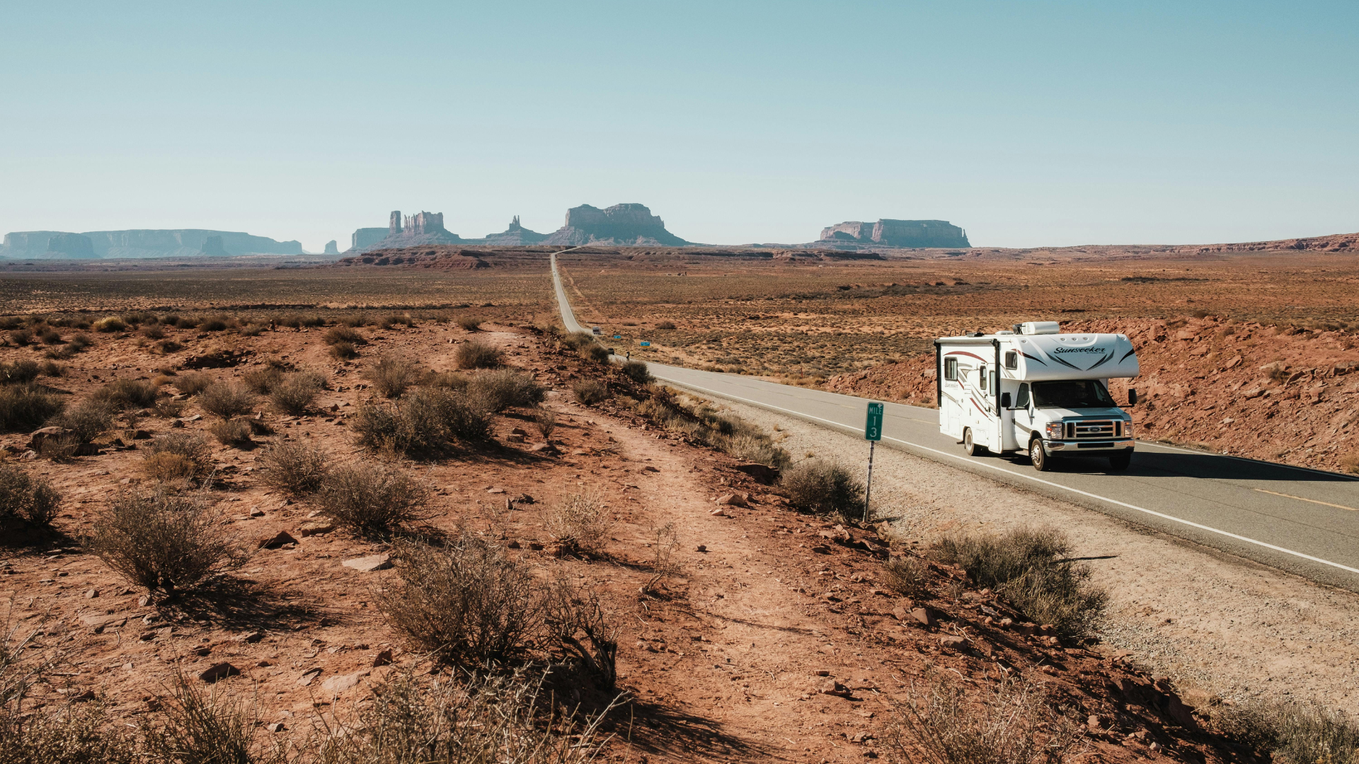RV driving down a road in a desert landscape with red rock formations in the background under a clear blue sky.