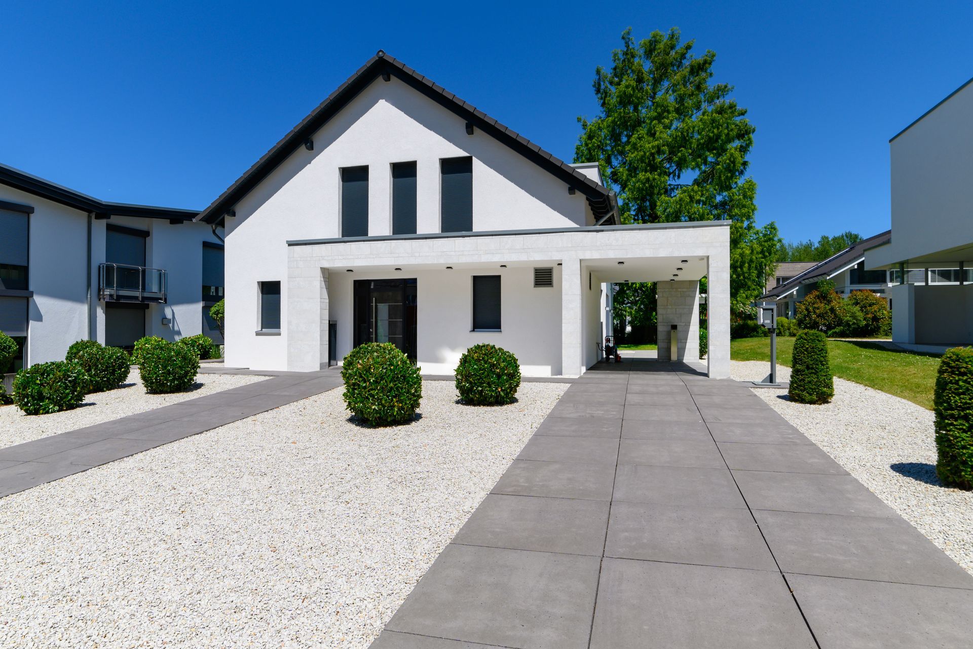 White modern house with carport, gray walkway, and landscaping, under blue sky.