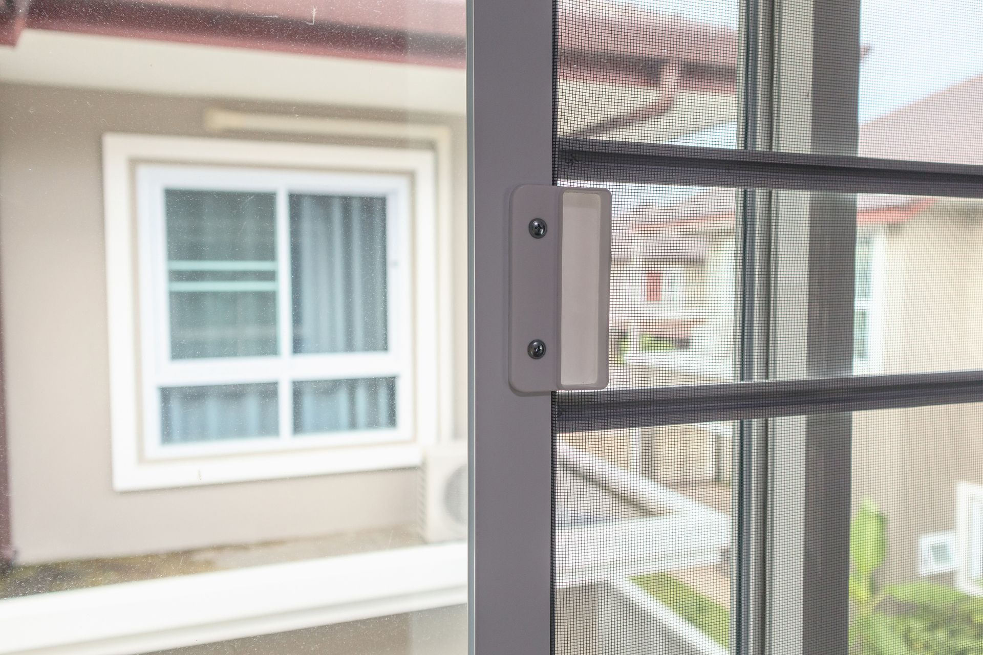 Close-up of a white screen door with a handle, with a window and a house in the background.