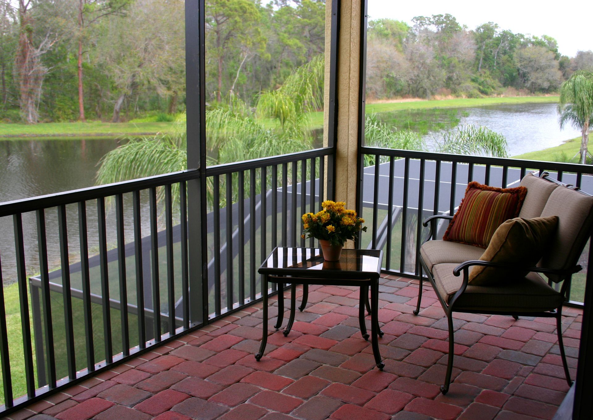Screened porch overlooking a lake, with a brick floor, wrought-iron furniture, and a vase of yellow flowers.