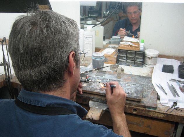 Man working on jewelry at a workbench, seen from the back. Mirror shows him working up close.