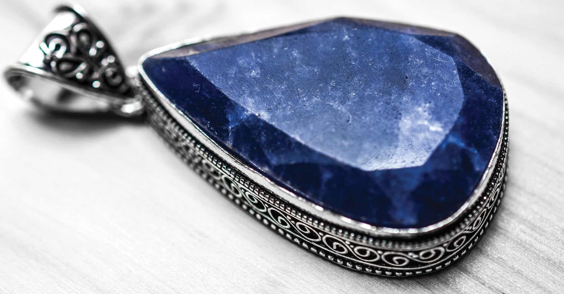 Close-up of a teardrop-shaped blue sapphire pendant with ornate silver metalwork on a light-colored surface.