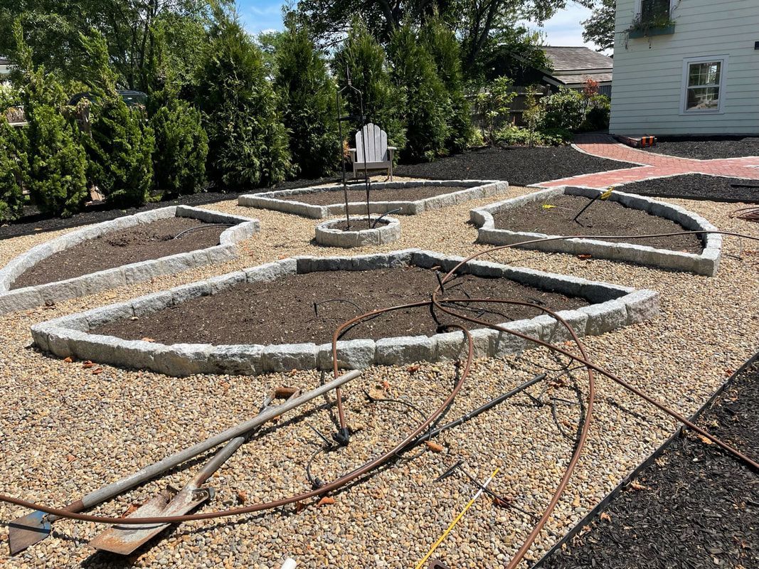 A garden with several raised beds outlined in gray stone, filled with soil, and surrounded by gravel.
